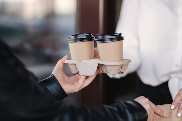 Kunden in Gesichtsmaske stehen im Freien und nehmen Paket mit Kaffee von Kassierer in Maske im Café nehmen