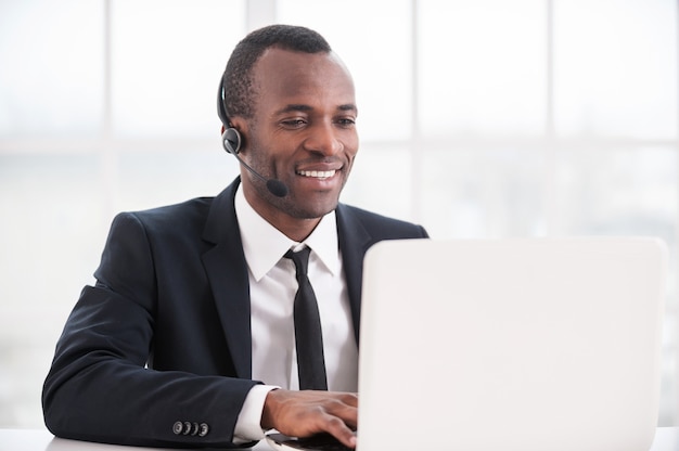 Kundendienstmitarbeiter bei der Arbeit. Fröhlicher junger Afrikaner in formeller Kleidung und Kopfhörer, der am Laptop arbeitet und lächelt