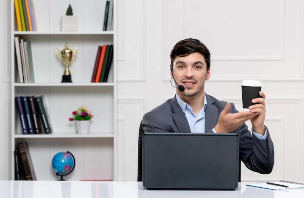Kundenservice süßer Kerl im grauen Anzug mit Computer und Headset, der eine Tasse hält und lächelt