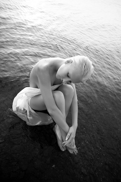Kunst nackte sexy Blondine mit kurzem Haarschnitt sitzt im Wasser am Ufer des Sees bei Sonnenuntergang. Nasses Haar und ein Frauenkörper. Abgeschiedener Strandurlaub. Schwarz und weiß