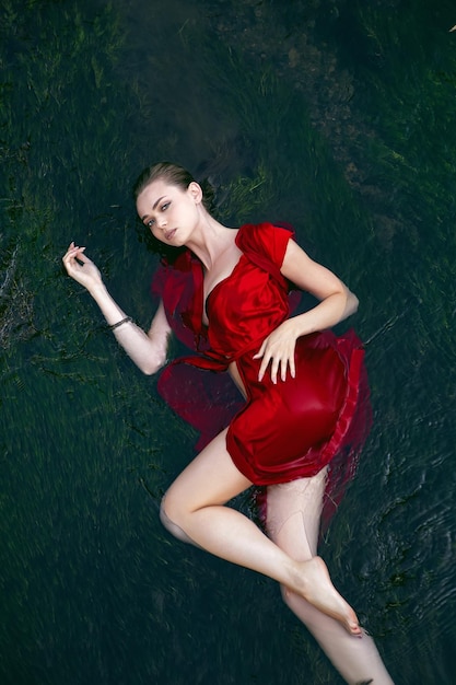 Foto kunstschönes romantisches porträt einer sexy jungen frau in einem roten kleid, das tagsüber in einem fluss mit grünalgen im sommer in der natur liegt