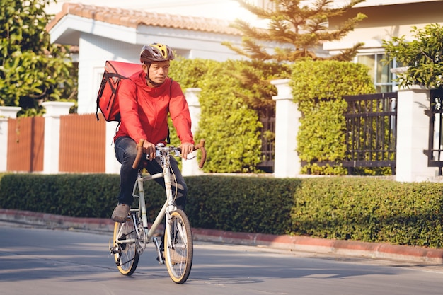 Kurier in roter Uniform mit einer Lieferbox auf dem Rücken, die Fahrrad fährt und auf das Handy schaut, um die Adresse zu überprüfen, um dem Kunden Lebensmittel zu liefern. Kurier auf einem Fahrrad liefert Essen in der Stadt.