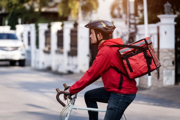 Kurier in roter Uniform mit einer Lieferbox auf dem Rücken, die Fahrrad fährt und auf das Handy schaut, um die Adresse zu überprüfen, um dem Kunden Lebensmittel zu liefern. Kurier auf einem Fahrrad liefert Essen in der Stadt.