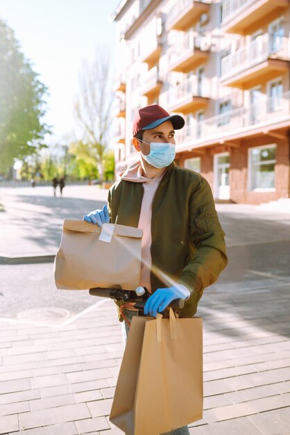 Kurier in Schutzmaske und medizinischen Handschuhen auf einem Roller liefert Bastelpapierpaket mit Lebensmitteln. Lieferservice unter Quarantäne, Krankheitsausbruch, Coronavirus-Covid-19-Pandemie.