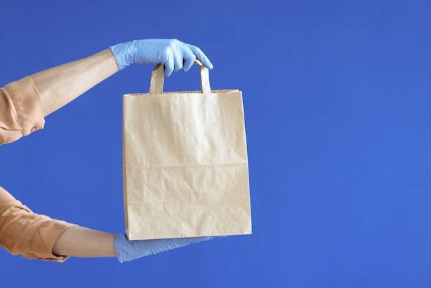 Kurierhände in Gummihandschuhen halten Papiertüte auf blauem Hintergrund