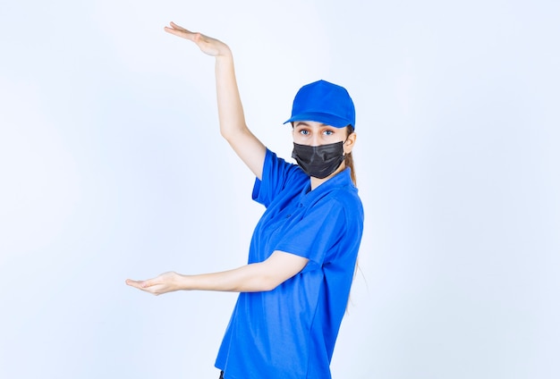 Kurierin in Maske und blauer Uniform, die die Höhe eines Objekts zeigt
