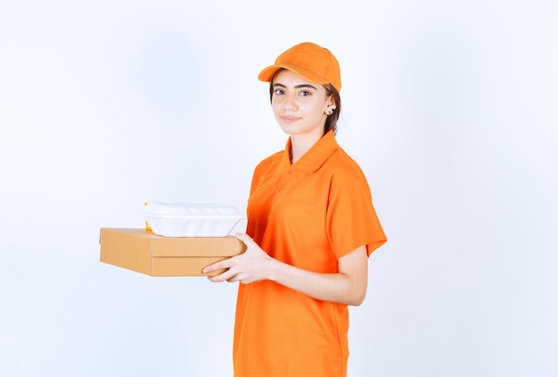 Kurierin in orangefarbener Uniform, die eine weiße Box zum Mitnehmen mit einem Papppaket hält