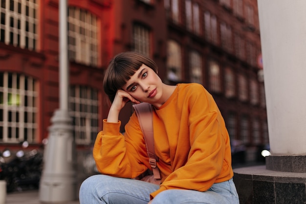 Kurzhaarige Frau im orangefarbenen Pullover sitzt draußen