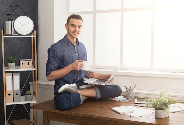 Kurzurlaub für Yoga im Büro. Flexibler Mann, der Yoga am Arbeitsplatz praktiziert, auf dem Schreibtisch in Lotuspose sitzt und Kaffee mit Laptop trinkt, Platz kopieren. Aktiver Mitarbeiter bei der Arbeit, gesundes Lebensstilkonzept