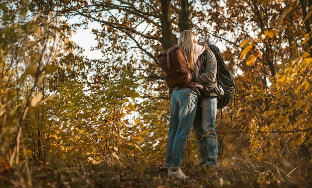 Kuss von verliebten Rucksacktouristen, die sich umarmen, während sie allein vor dem Hintergrund des Herbstwaldes im Freien stehen