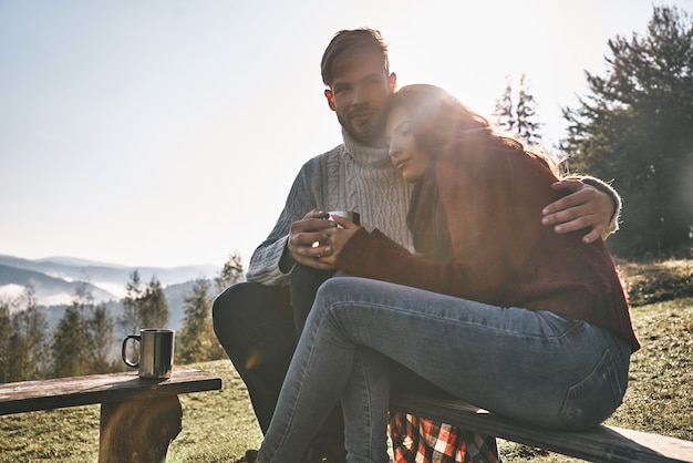 Él siempre está ahí para ella. Hermosa joven pareja tomando café por la mañana mientras está sentado en el banco en las montañas