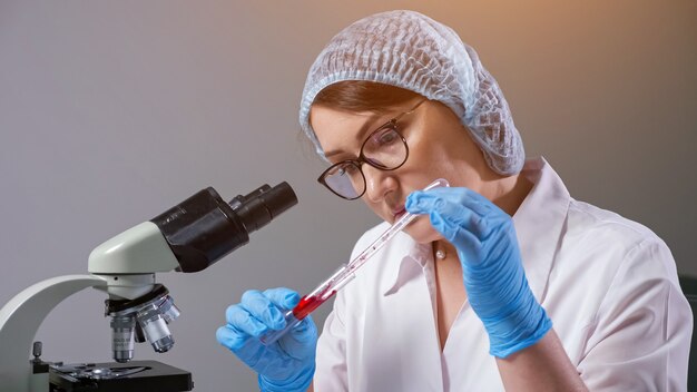 Laborassistentin mit Brille mischt Blut und transparente Flüssigkeit im Reagenzglas in der Nähe des Mikroskops, das in einem modernen Krankenhaus in der Nähe arbeitet