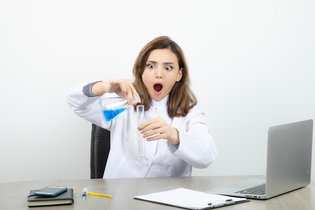 Laborforscherin, die am schreibtisch sitzt und medizinische glasflasche mit blauer flüssigkeit hält. Foto in hoher Qualität