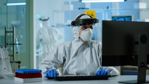 Labortechnikerassistent im PPE-Anzug, der die Blutprobe in der Reagenzglastypisierung am PC analysiert. Arzt, der mit verschiedenen Bakterien, Geweben, pharmazeutischer Forschung für Antibiotika gegen Covid19 arbeitet.