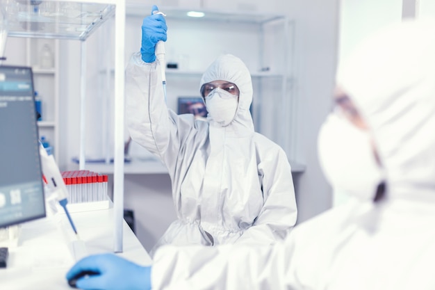 Laborwissenschaftler im Gesundheitswesen, der eine Mikropipette mit einer Probe in einem PPE-Anzug gegen Coronavirus hält. Team von Mikrobiologen im Forschungslabor, das Experimente während der globalen Pandemie durchführt