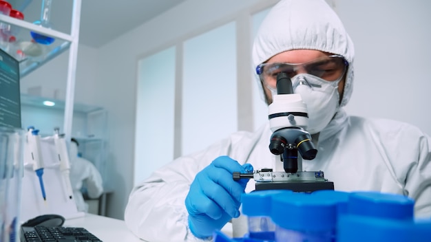Laborwissenschaftler im PPE-Anzug, der Experimente durchführt, die im modernen Labor durch ein Mikroskop schauen. Mann, der mit verschiedenen Bakteriengeweben, Blutproben, Konzept der medizinischen Forschung für Antibiotika arbeitet