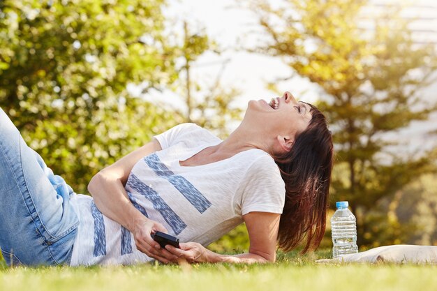 Lachende Frau, die draußen im Gras mit Handy liegt