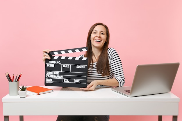 Lachende Frau mit klassischer schwarzer Filmklappe, die an einem Projekt arbeitet, während sie im Büro mit Laptop sitzt, isoliert auf pastellrosa Hintergrund. Erfolgsgeschäftskarrierekonzept. Platz kopieren.