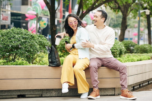 Lachende junge Chinesin versteckt Smartphone vor ihrem Freund, wenn sie auf der Bank im Park sitzt