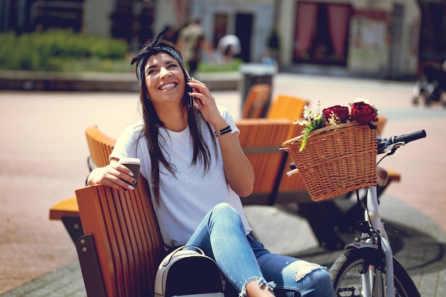 Lachende junge Frau, die auf dem Smartphone spricht, Kaffeepause macht, an einem sonnigen Tag auf einer Bank sitzt, neben dem Fahrrad mit Blumenkorb.