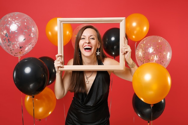 Lachende junge Frau im kleinen schwarzen Kleid feiert, Bilderrahmen auf leuchtend roten Hintergrundluftballons halten. Valentinstag, Frauentag, Happy New Year Geburtstag Mockup Urlaub Party Konzept.