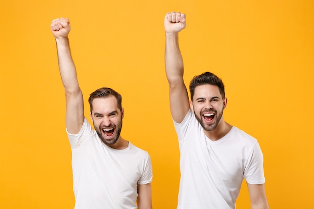 Lachende junge Männer Jungs Freunde in weißen leeren leeren T-Shirts posieren isoliert auf gelb orange Wand in. Menschen Lifestyle-Konzept. Steigende Hände hoch mit Siegergeste.