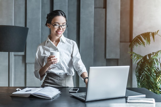 Lachende junge Unternehmerin, die eine Tasse Kaffee trinkt und in ihrem Heimbüro am Laptop arbeitet