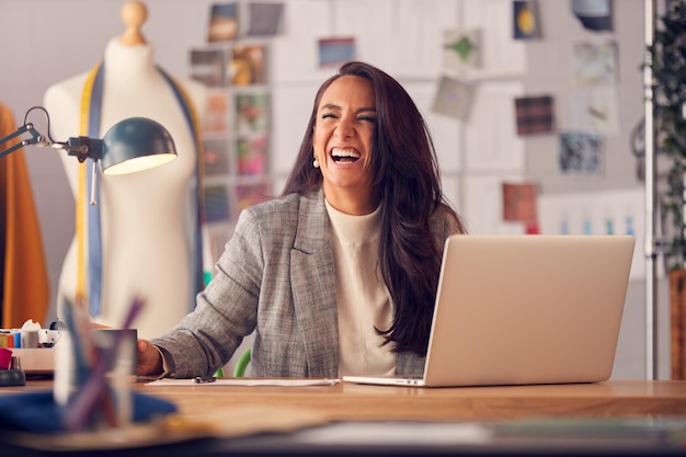 Lachende Modedesignerin im Studio, die an Skizzen oder Dokumenten am Schreibtisch mit Laptop arbeitet