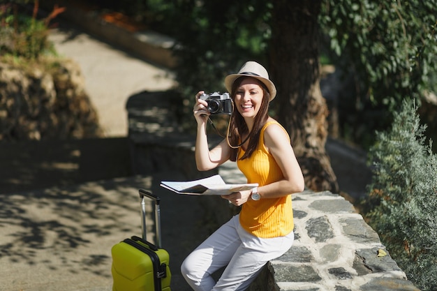 Lachende Reisende Touristenfrau in Hut mit Kofferstadtplan fotografieren auf Retro-Vintage-Fotokamera in der Stadt im Freien. Mädchen, das ins Ausland reist, um am Wochenende zu reisen. Tourismus Reise Lebensstil.