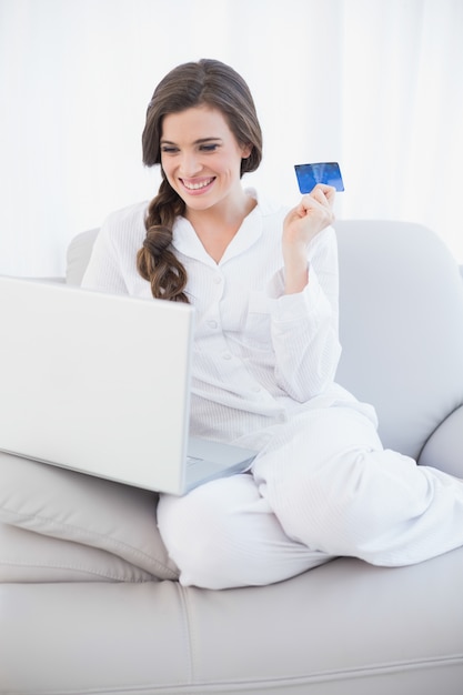 Lachende zufällige braune behaarte Frau in den weißen Pyjamas, die online mit ihrem Laptop kaufen
