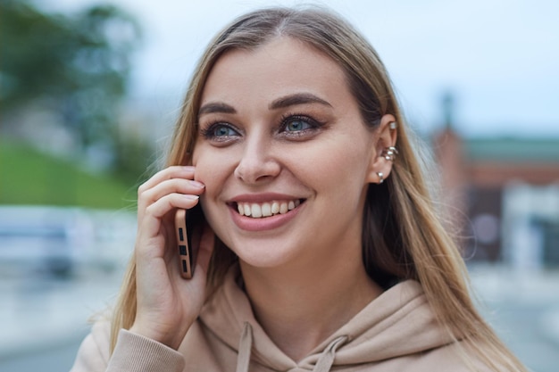 Lachender blonder Teenager in einem beigen Sweatshirt, der auf einem Handy spricht