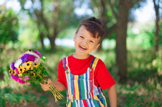 Lachender Junge mit einem Blumenstrauß der wilden Blumen in seinen Händen vor dem Hintergrund des Muttertags der Natur