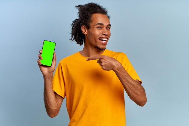 Lachender junger afrikanisch-amerikanischer Hipster-Mann zeigt auf modernen Smartphone-Greenscreen mit Alphakanal, isoliert auf blauem Hintergrund. Greenscreen-Werbefläche. Gadgets und Kommunikationskonzept