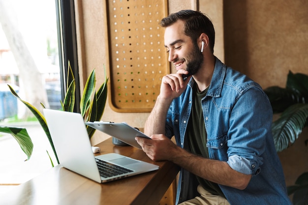 lachender kaukasischer Mann mit Jeanshemd mit Ohrstöpsel und Zwischenablage mit Laptop, während er im Café drinnen arbeitet