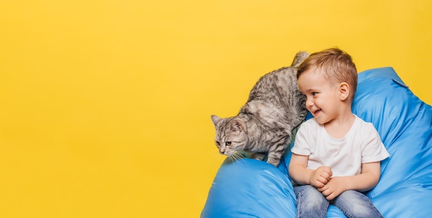 Lachender kleiner Junge an einer gelben Wand sitzt mit einer Katze, die auf einem blauen Stuhl sitzt