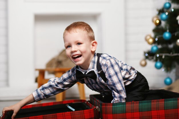 Lachender kleiner Junge spielt versteckt in einem roten karierten Koffer im Innenraum mit Weihnachtsdekorationen