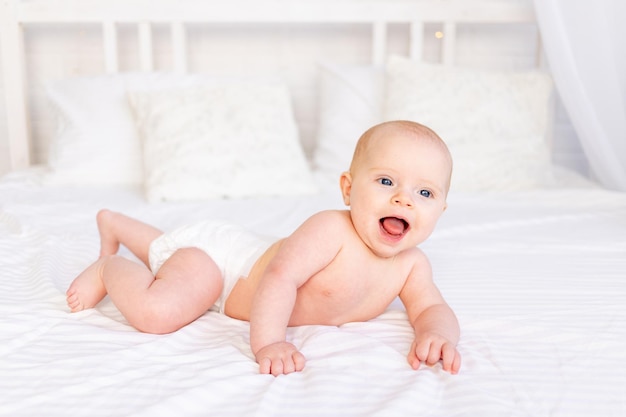 Lachendes Babymädchen in Windeln in einer Krippe auf einem weißen Baumwollbett, das auf dem Bauch im Kinderzimmer liegt, lächelndes Neugeborenes am Morgen