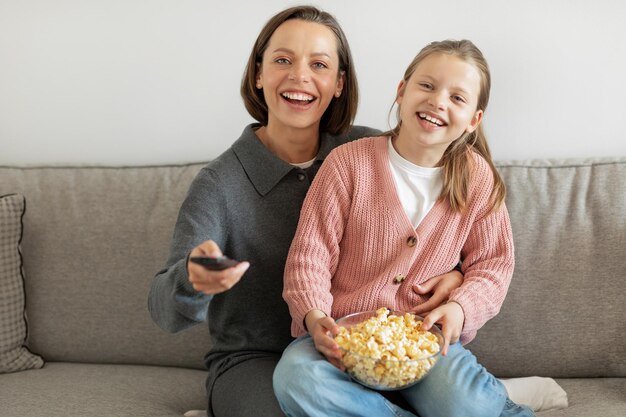 Lachendes, glückliches europäisches Teenager-Mädchen und tausendjährige Mutter mit Fernbedienung, die Popcorn beim Fernsehen isst