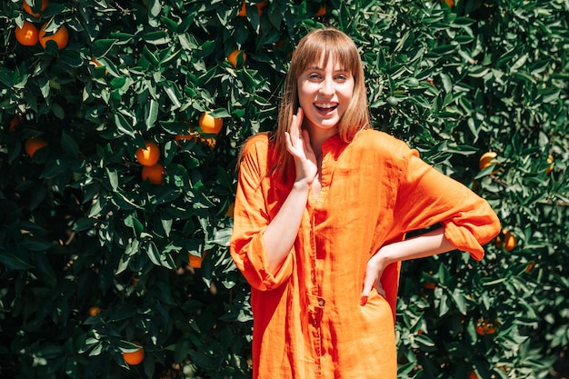 Lachendes junges Mädchen in orangefarbenem Kleid schaut in die Kamera, indem es die Hand auf die Taille im orangefarbenen Garten legt