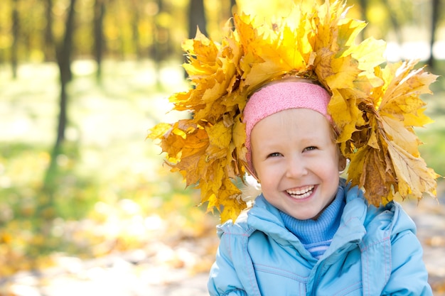 Lachendes kleines Mädchen mit einem Hut aus gelbem Herbstlaub, den sie selbst gesammelt hat