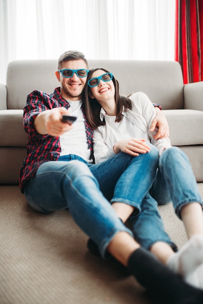 Lachendes Paar in 3D-Brille sitzt auf dem Boden gegen Couch und sieht zu Hause fern