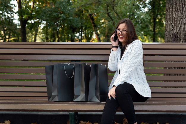 Lachendes schönes Frauengespräch am Telefon. Ruhe im Park mit Einkaufstüten. Glückliche junge Frau nach dem Einkaufen.