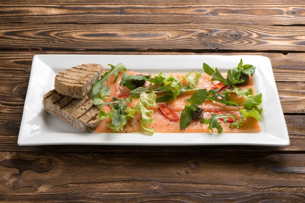 Lachs- oder Forellen-Carpaccio mit Rucola, Paprika, Salat, Basilikum und gegrilltem Ciabatta in einer weißen Keramikplatte auf dem hölzernen Küchentisch.