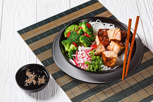 Lachs-Teriyaki mit Reisnudeln, Brokkoli, Rettich und Frühlingszwiebeln in einer Schüssel mit Essstäbchen auf einem weißen Holztisch auf einer Bambusmatte