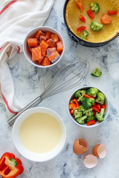 Lachsfilet, Brokkoli und Paprika in einer Schüssel, Milch und Eier mischen, Teig auf einem weißen Tisch säuern