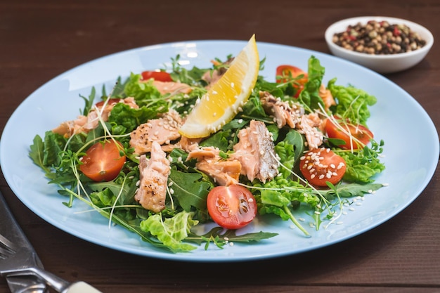 Lachssalat mit grünen Blättern Tomaten-Zitronen-Sesam in blauer Platte auf Holztisch