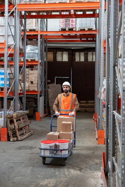 Lader in Arbeitskleidung, der Wagen mit Waren schiebt, während er an Regalen entlang geht