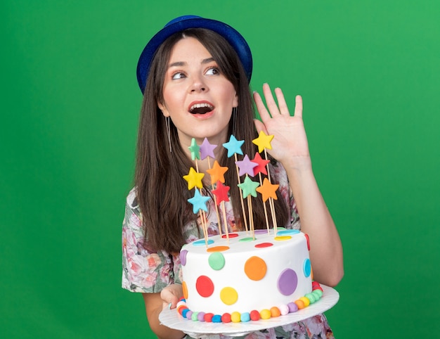 Lado alegre jovem com um chapéu de festa segurando um bolo e mostrando um gesto de olá isolado na parede verde