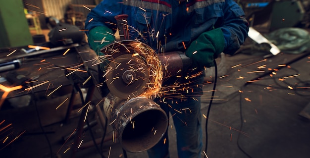 Lado close-up vista de homem trabalhador focado profissional em uniforme corta escultura de tubo de metal com um grande moedor elétrico enquanto faíscas voam na oficina de tecido industrial.