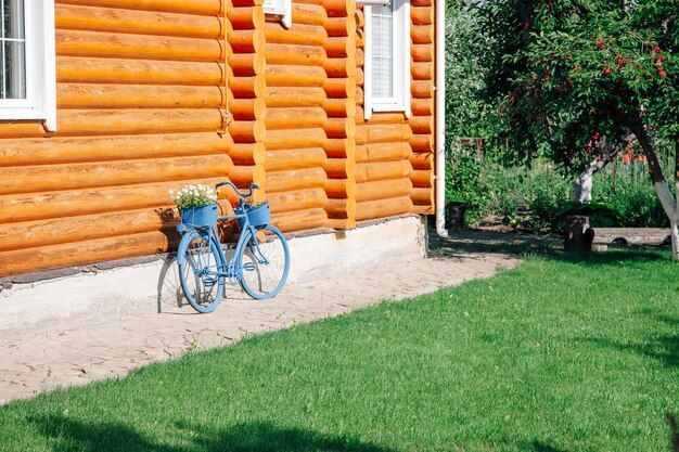 Lado ensolarado da casa de campo de madeira com bicicleta azul decorativa com vaso de flores no porta-malas apoiado contra a fachada da casa à esquerda e cerejeira à direita durante o dia na vida da vila de verão